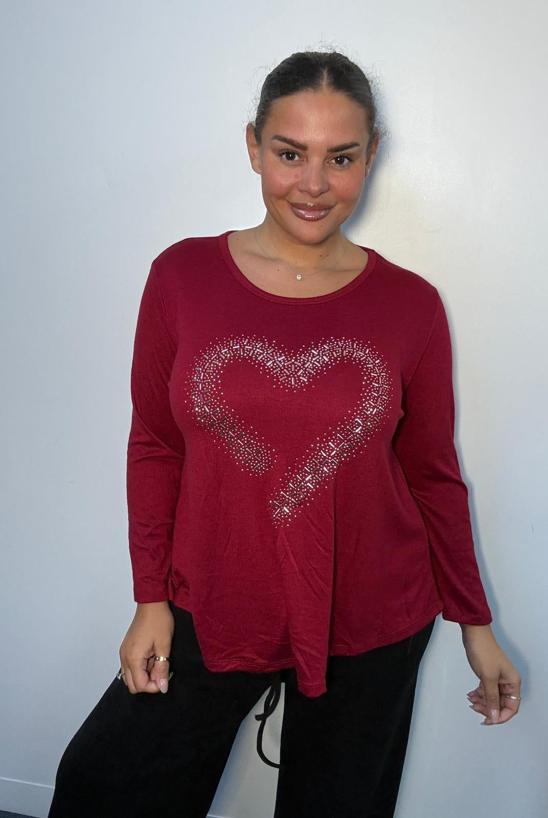 Plus-size model wearing a burgundy long-sleeve top featuring a sparkling diamante-style heart design and asymmetric pointed hem.