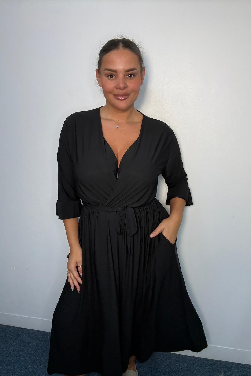 Plus-size model wearing a black wrap-style midi dress with V-neckline, matching tie waist, three-quarter flared sleeves and flowing skirt.