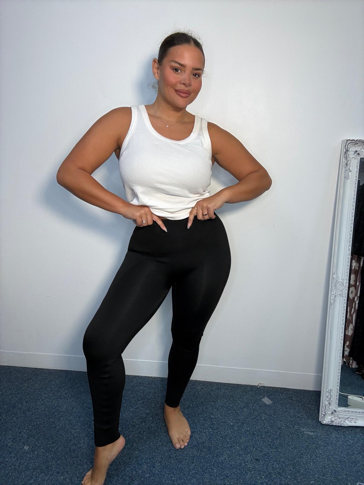 Plus-size model wearing black high-waisted fitted leggings with a white sleeveless vest top, showing the smooth waistband and full-length fit.
