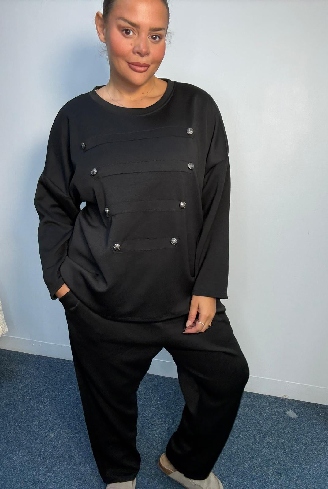 Plus-size model wearing a black two-piece lounge set featuring a long-sleeve top with silver-tone button detailing and matching relaxed-fit trousers.