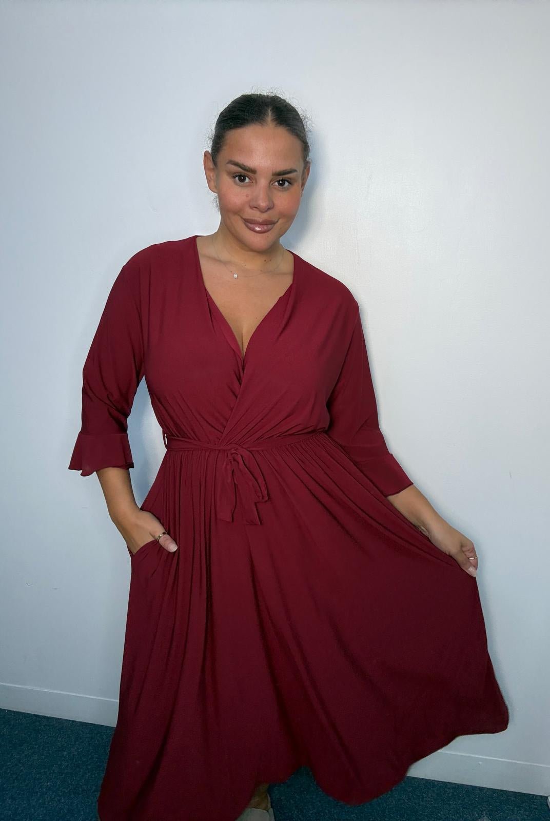 Plus-size model wearing a burgundy wrap-style midi dress with V-neckline, matching tie waist, three-quarter flared sleeves and flowing skirt.