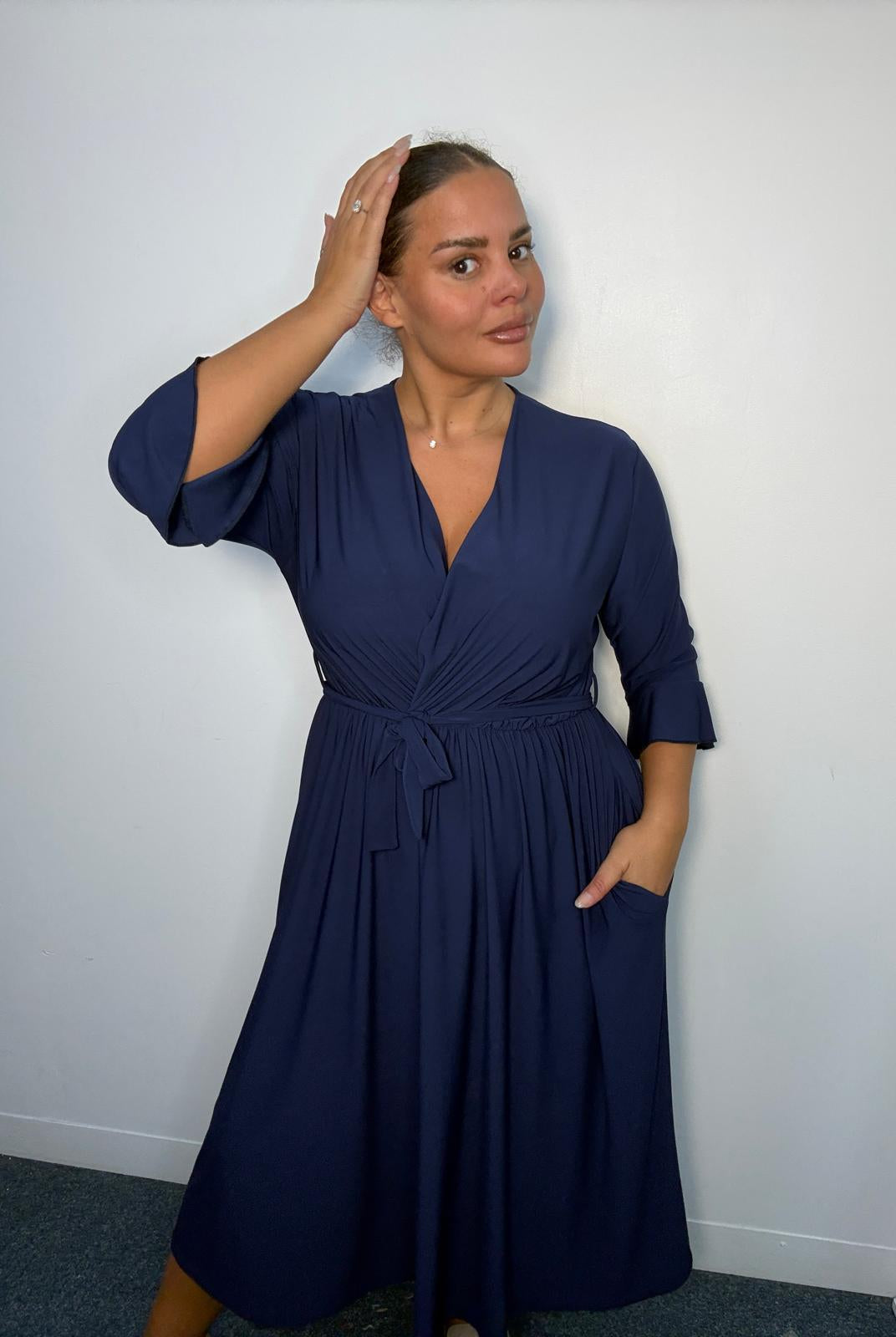 Plus-size model wearing a navy wrap-style midi dress with V-neckline, matching tie waist, three-quarter flared sleeves and flowing skirt.