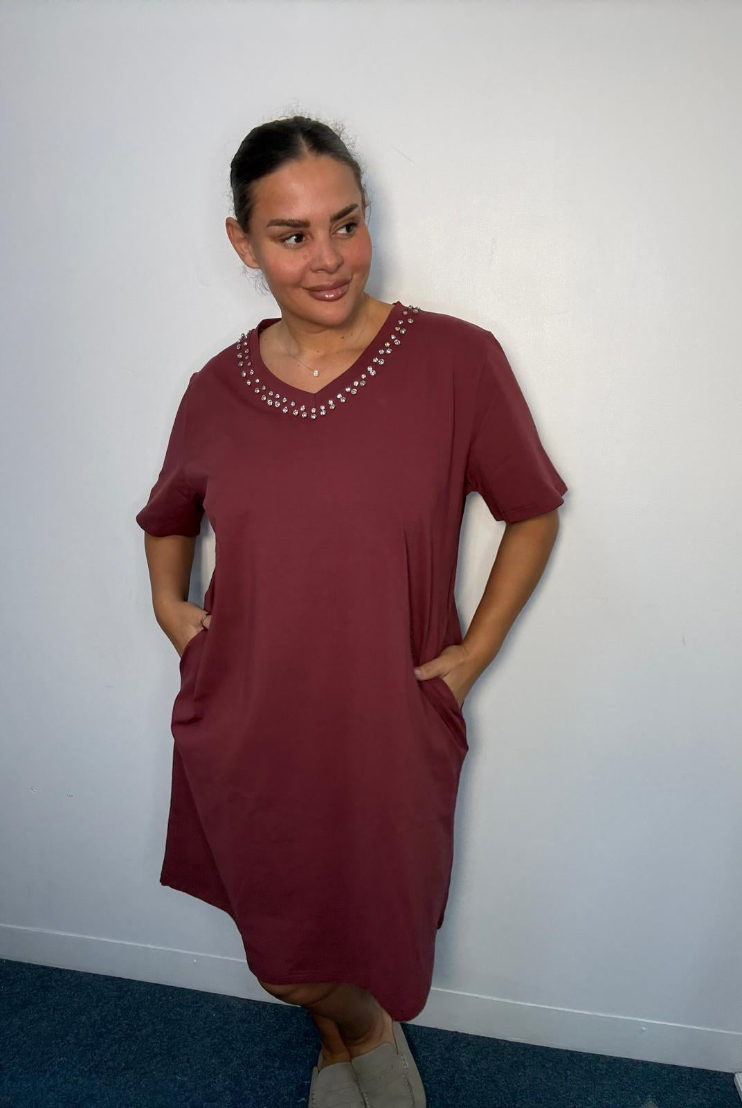 Plus-size model wearing a burgundy midi dress with diamante-style V-neck detailing and hands in the side pockets.