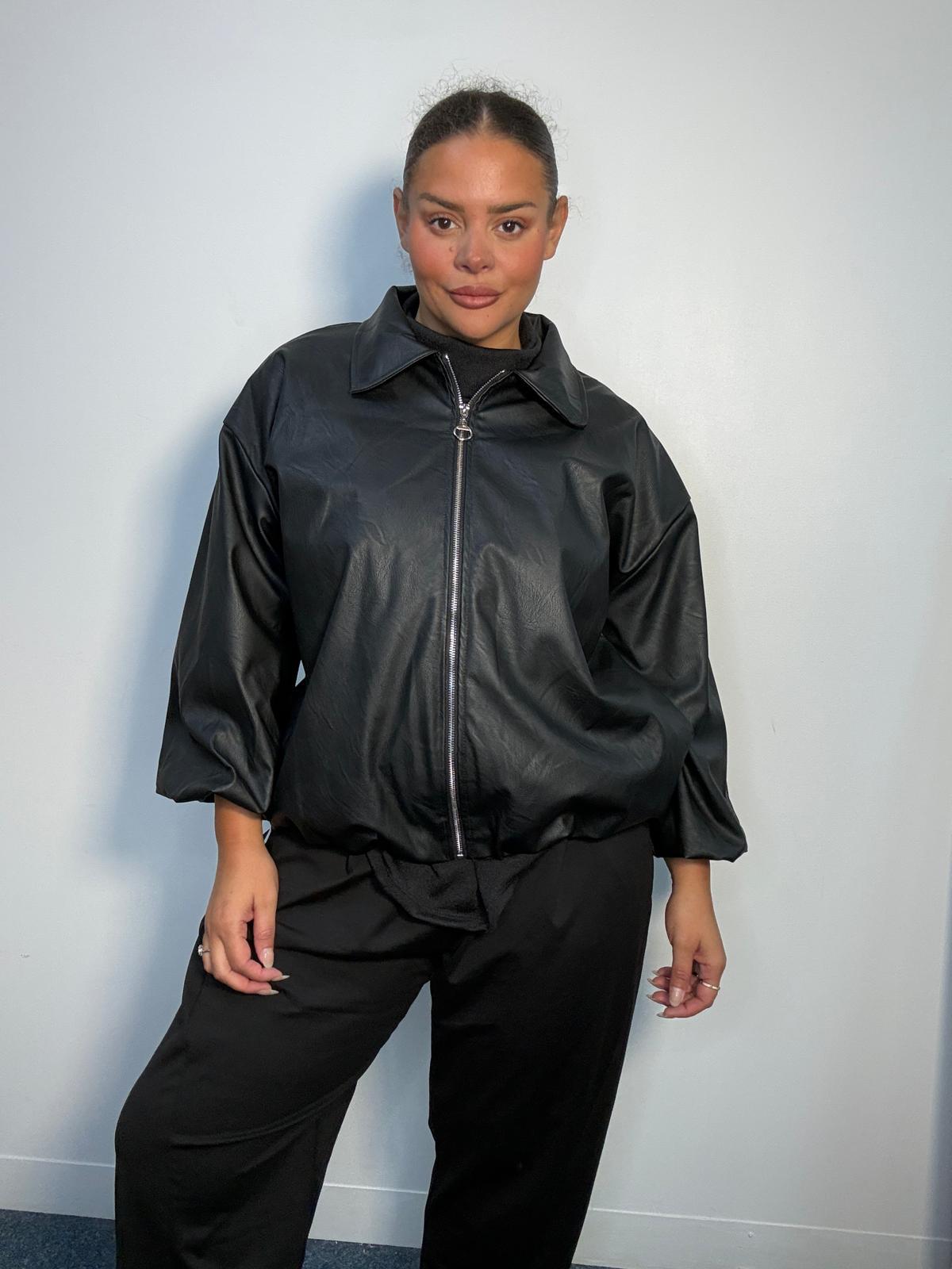Plus-size model wearing a black faux leather-look bomber jacket with a silver front zip, pointed collar and gathered sleeves.