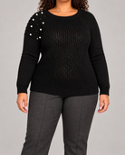 A woman wearing a black ribbed knit sweater with pearl embellishments on one shoulder, paired with dark gray trousers, against a solid gray studio background.