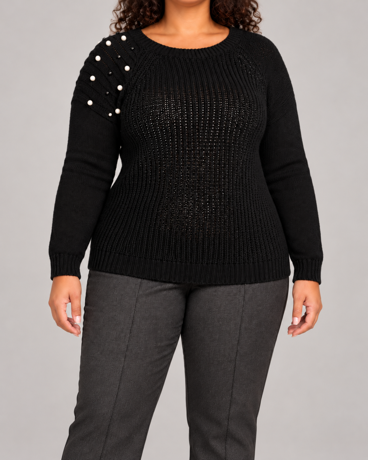 A woman wearing a black ribbed knit sweater with pearl embellishments on one shoulder, paired with dark gray trousers, against a solid gray studio background.