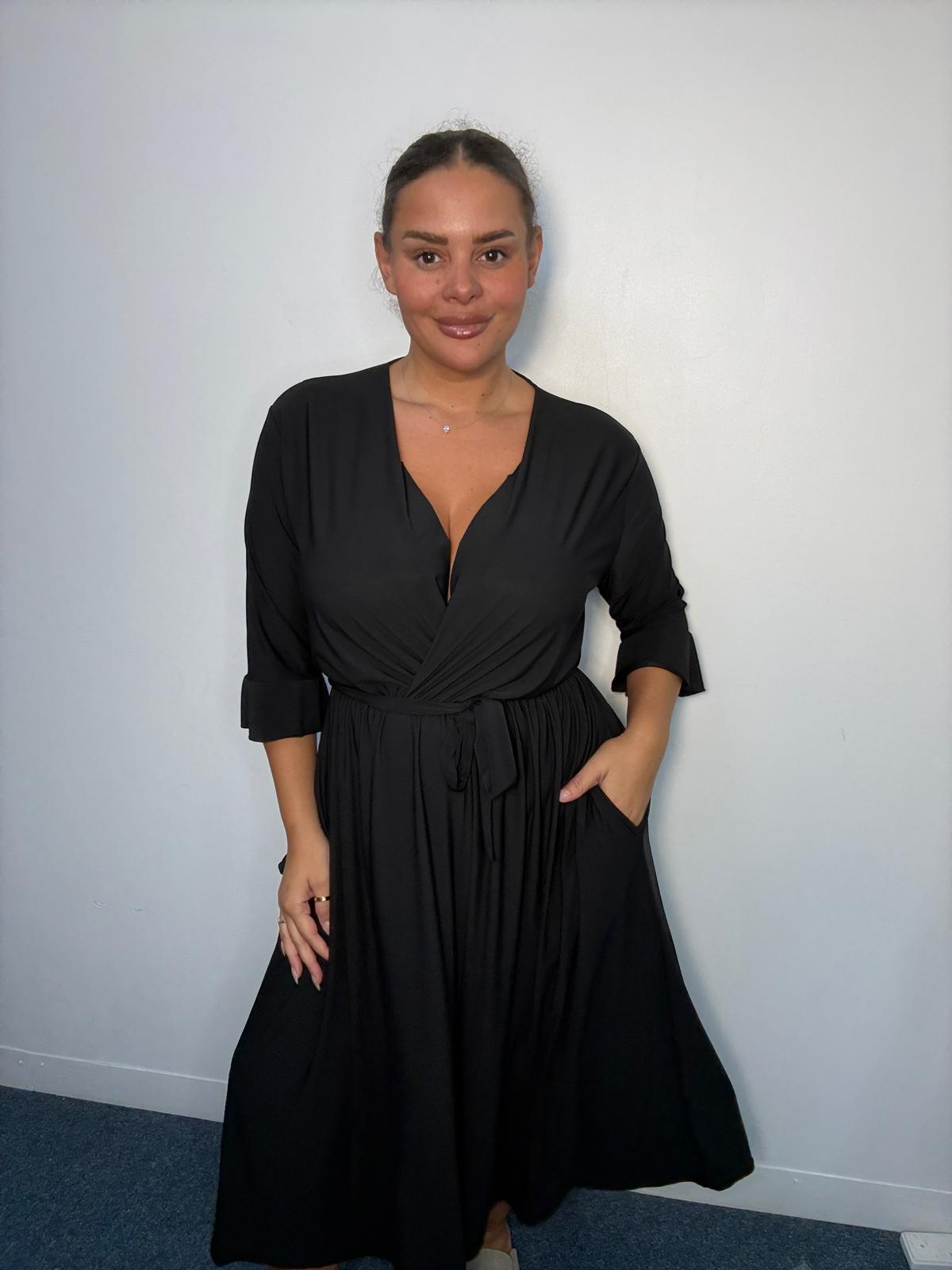 Plus-size model wearing a black wrap-style midi dress with V-neckline, matching tie waist, three-quarter flared sleeves and flowing skirt.