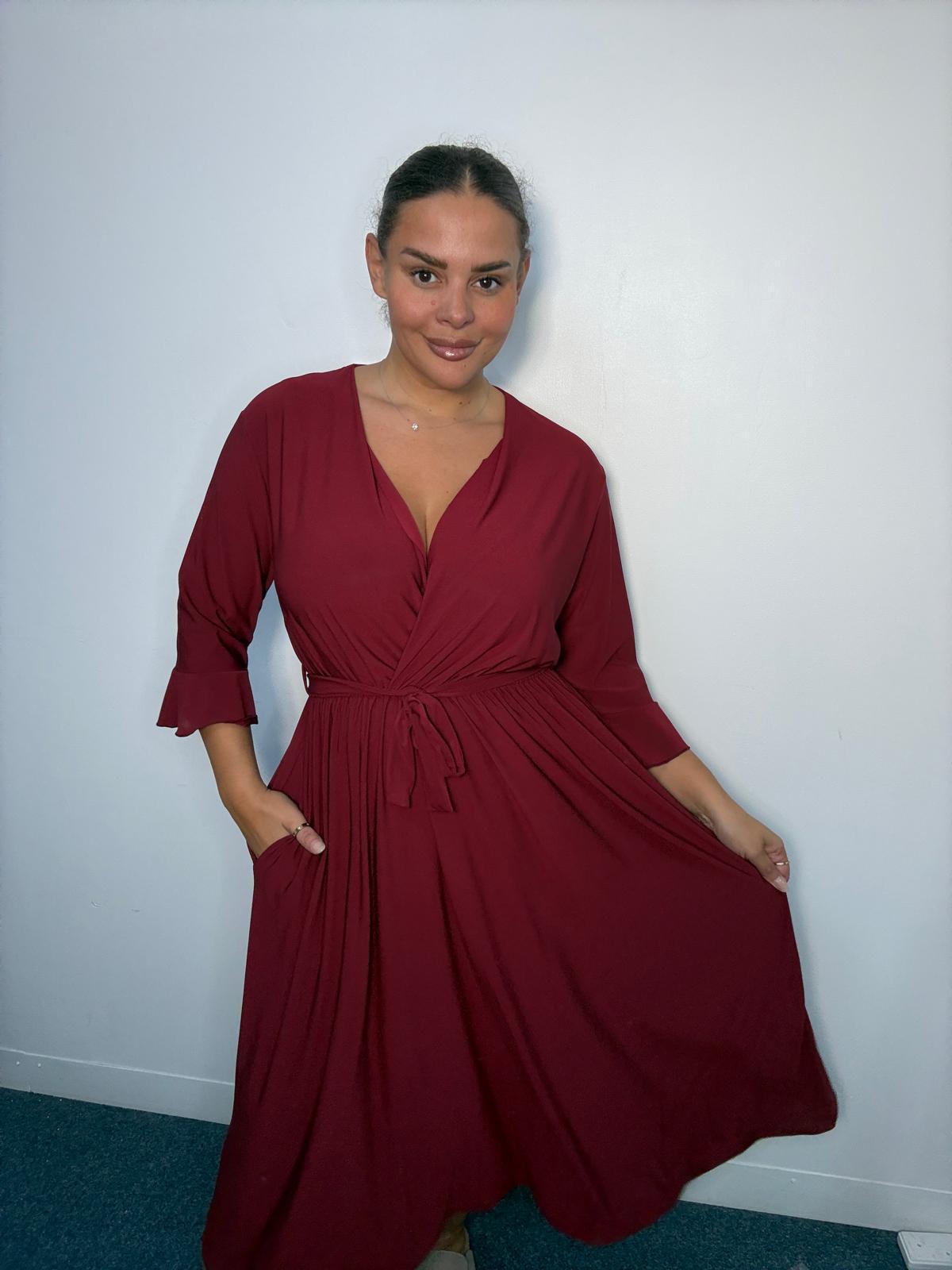 Plus-size model wearing a burgundy wrap-style midi dress with V-neckline, matching tie waist, three-quarter flared sleeves and flowing skirt.