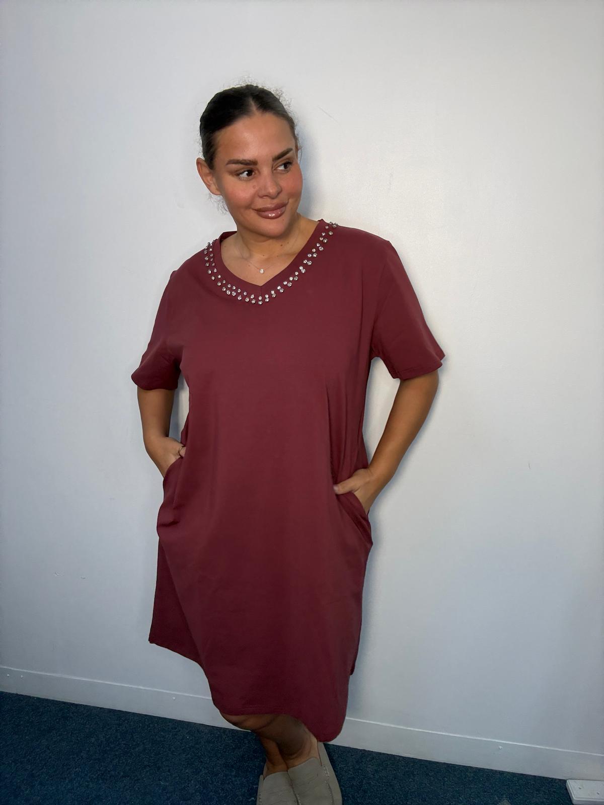 Plus-size model wearing a burgundy midi dress with diamante-style V-neck detailing and hands in the side pockets.